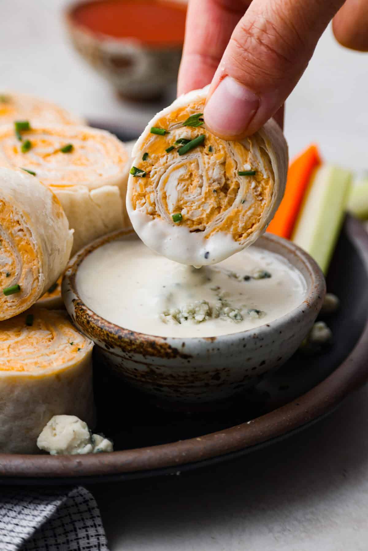 These zesty Buffalo Chicken Pinwheels are an easy snack or appetizer that everyone will devour! A creamy buffalo-flavored spread full of shredded chicken, seasonings, and cheeses all rolled inside of a soft tortilla. Slice into cute pinwheels and enjoy! Close view of a pinwheel dipping into blue cheese dressing.