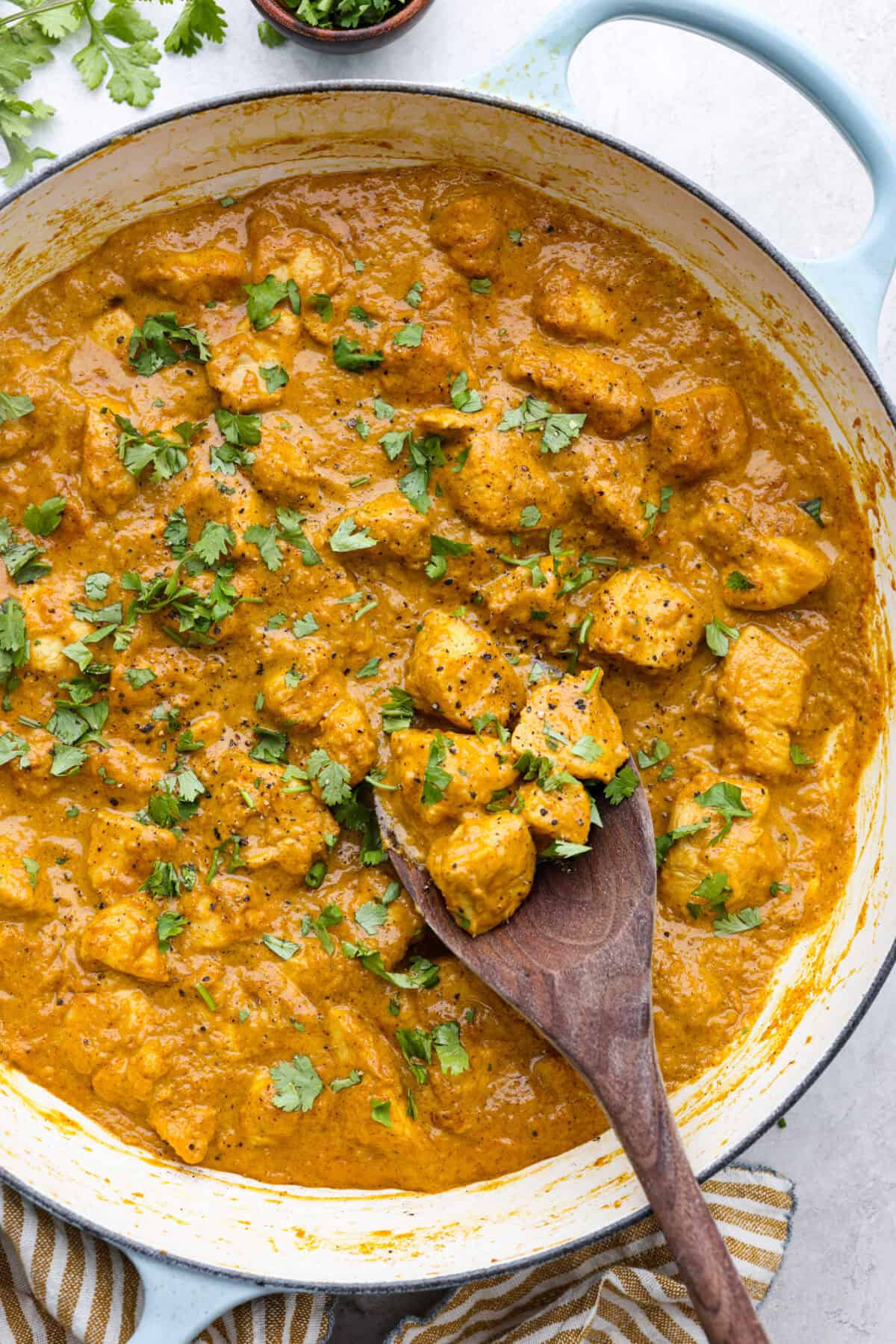 Chicken korma is a traditional Indian chicken dish. It's made with tender chicken smothered in a creamy, wonderfully seasoned sauce. It's so good your family will think you got takeout! Overhead shot of chicken korma in a skillet with wooden spoon and garnished with cilantro.