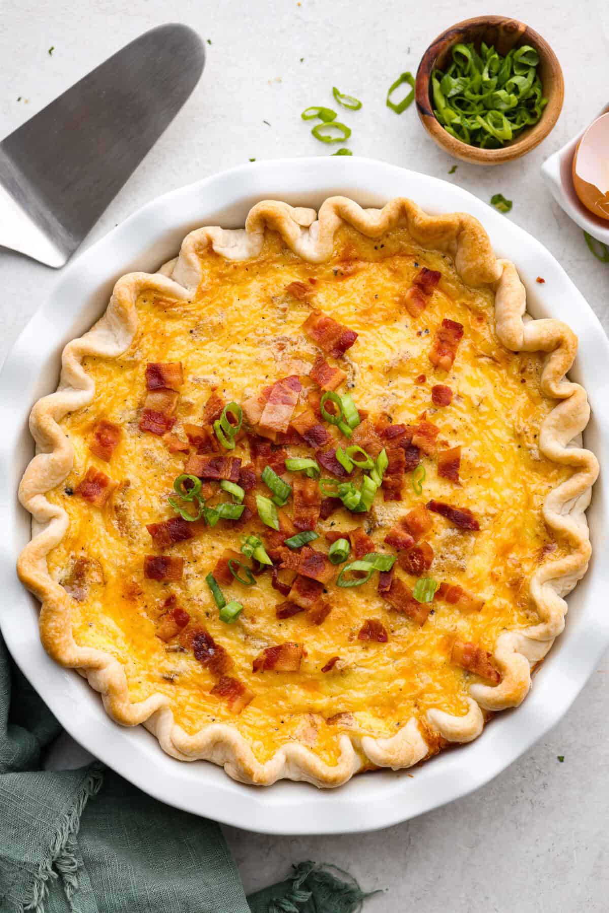 Cowboy Quiche is the ultimate hearty, satisfying breakfast that brings classic comfort to the table. It is filled with smoky bacon, caramelized onions, and cheddar cheese, all baked together in a buttery pie crust. It’s rich, savory, and packed with bold flavor. Overhead shot of baked cowboy quiche with green onion and bacon bits as garnish on the top.