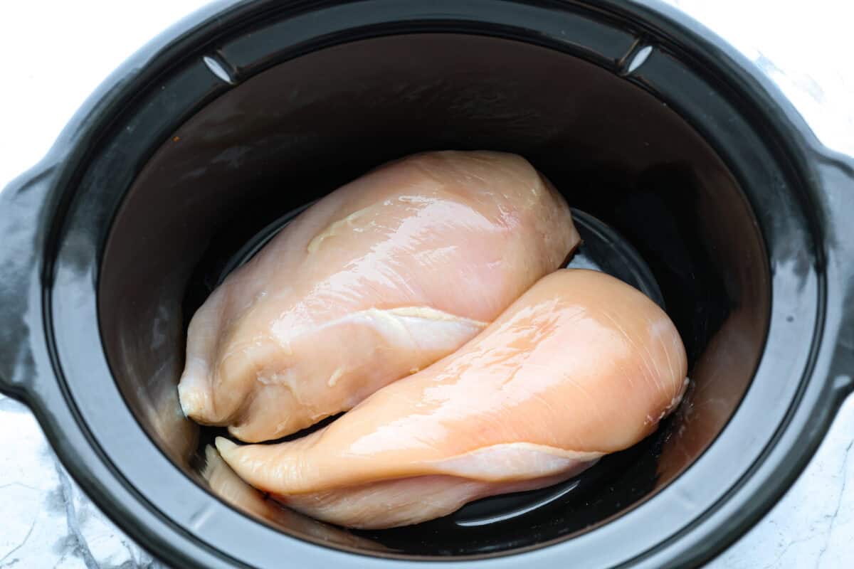This crockpot chicken alfredo is a family dinner that's packed with flavor! The juicy, tender chicken cooked with creamy, cheesy pasta simply can't be beat! Plus, it’s super easy when your crock pot does all the work! Overhead shot of raw chicken in the bottom of the crockpot.