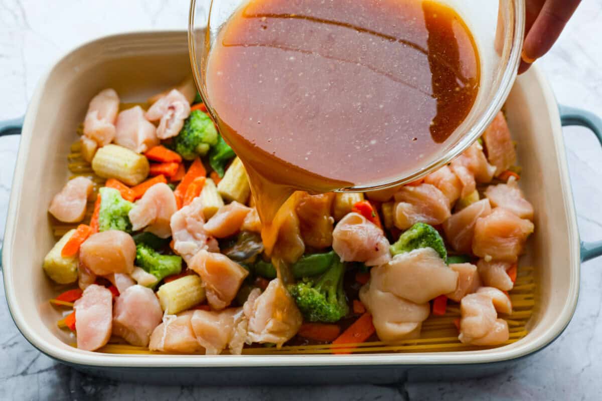 Honey Sesame Chicken Noodle Bake is the perfect dinner solution when you need something incredibly tasty and totally fuss-free! This is a complete meal with delicious Asian flavors in one pan, without needing to pre-cook anything. Overhead shot of someone pouring the sauce over the chicken, veggies and noodles in a baking dish.