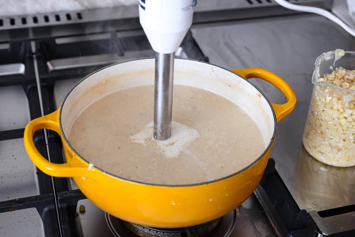 Mexican street corn soup has all the incredible flavors of street corn in a warm, comforting, delicious soup. This soup has fresh corn, lots of cotija cheese, spices, cilantro, lime juice, and sour cream to make it smooth and creamy. Angle shot of someone blending the soup with an immersion blender in the pot with a measuring cup with corn next to it.