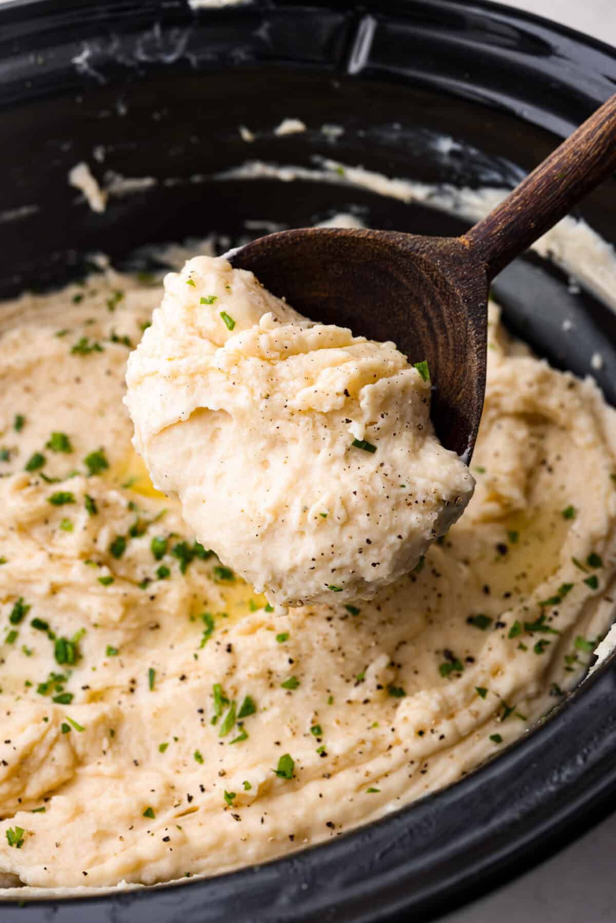 Million dollar mashed potatoes are unmatched when it comes to creamy velvety mashed potatoes. They get their intense flavors and smooth texture from letting the potatoes slow cook in chicken broth then mixing them with cream cheese, butter and garlic. Close up shot of someone scooping a serving of million dollar mashed potatoes out of the crock pot.