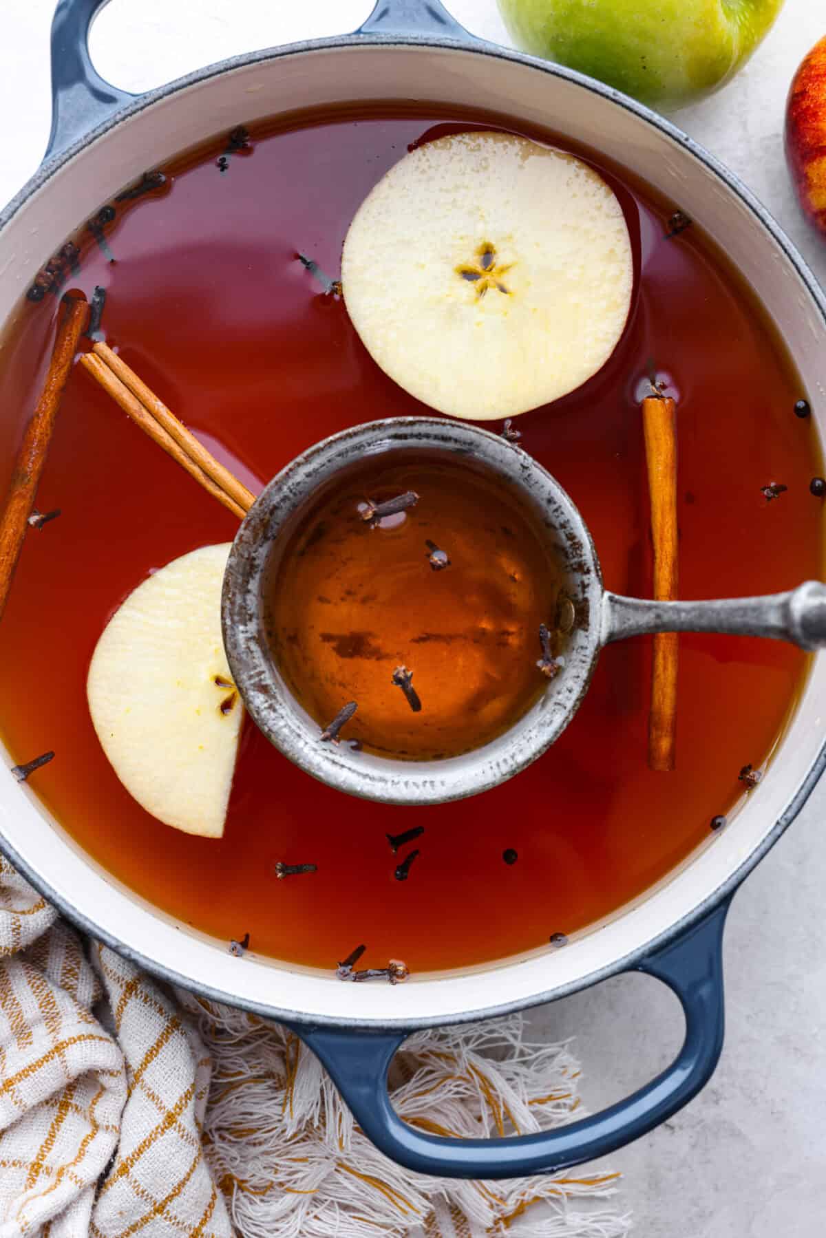 Mulled cider is a warm, inviting drink that is mixed with citrus and spices. Take your apple cider game up a notch with this easy recipe. Your house will smell amazing as well! Overhead shot of mulled cider with apple slices, cinnamon sticks cloves and allspice berries in a pot.