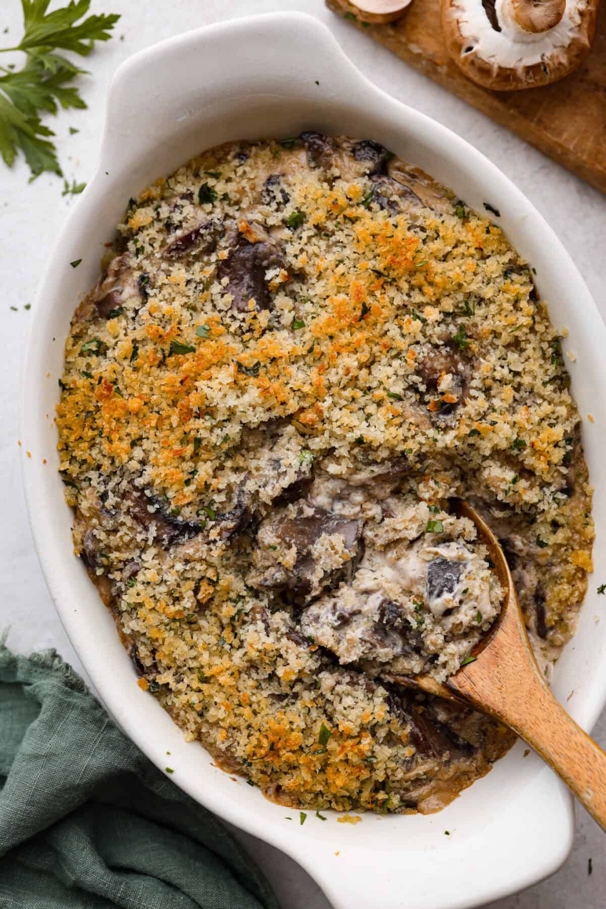 This creamy Mushroom Casserole is an impressive side dish to any meal! It's loaded with tender mushrooms mixed in a cheesy decadent sauce, then baked with a panko herb topping. Top view of mushroom casserole in a baking dish with a wood serving spoon.