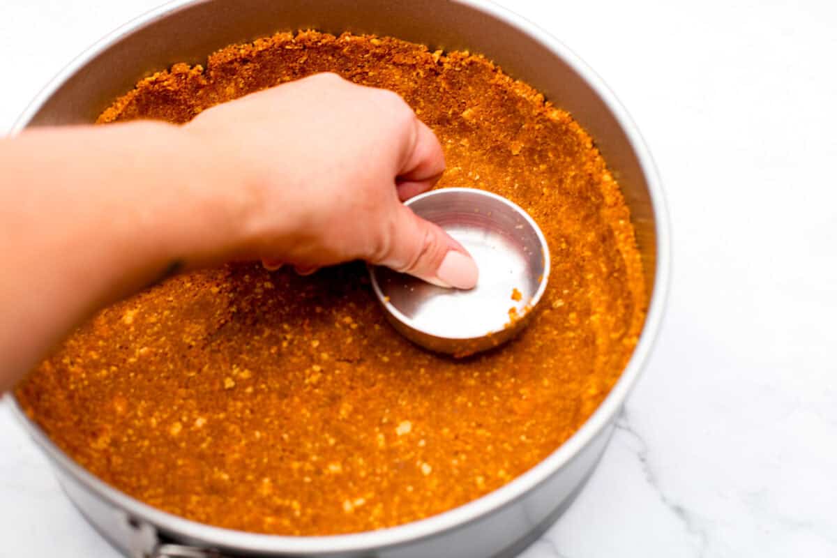 Delicious no-bake pumpkin cheesecake topped off with whipped cream is the perfect fall dessert. It has a creamy cheesecake filling with real pumpkin puree that sits in an irresistible Biscoff cookie crust.ย Itโs easy to make, rich, and perfectly spiced! Overhead shot of someone smashing the crust mix into the bottom of a spring form pan.