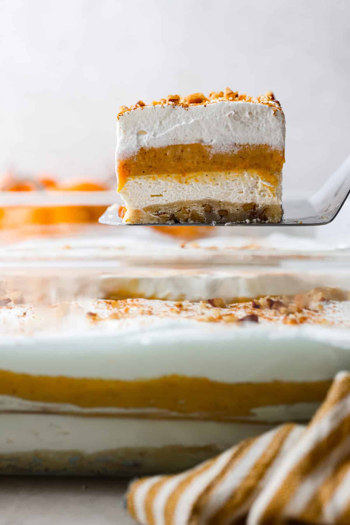 Fall in love with this Pumpkin Delight! A shortbread crust topped with a fluffy cheesecake mixture, a creamy pumpkin layer, and whipped topping. Every bite of this irresistible layered dessert is pumpkin heaven! Close view of a slice of pumpkin delight lifting from the pan.