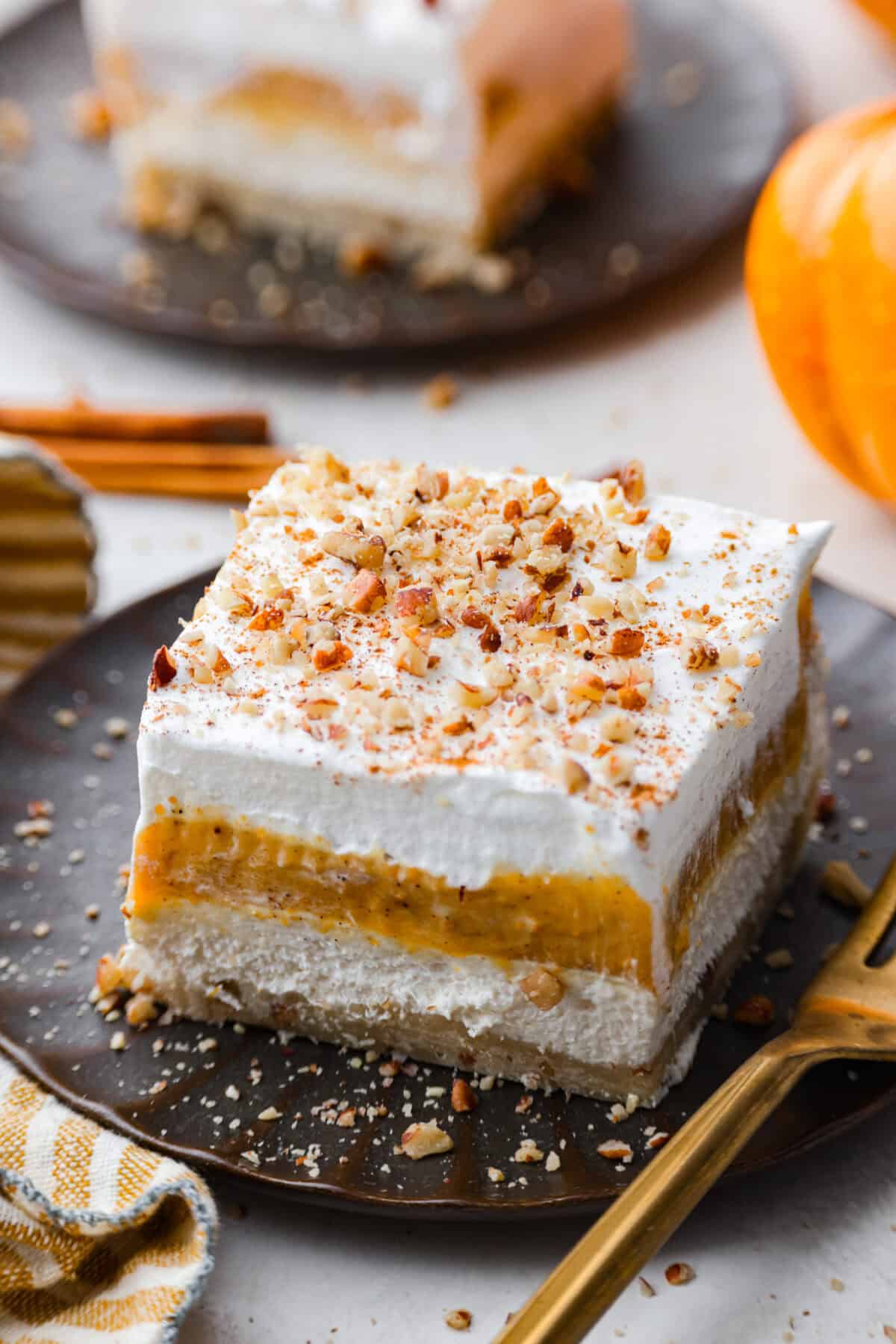 Fall in love with this Pumpkin Delight! A shortbread crust topped with a fluffy cheesecake mixture, a creamy pumpkin layer, and whipped topping. Every bite of this irresistible layered dessert is pumpkin heaven! Close view of a slice of pumpkin delight on a plate with a fork.