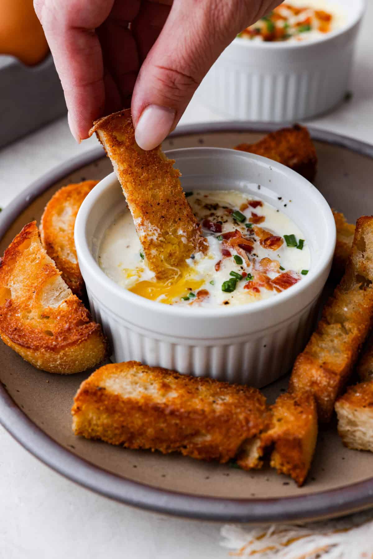 Shirred Eggs are a breakfast worth waking up for in the morning! They're a creamy, custardy combination of eggs, cheese, cream, and bacon that bake in individual ramekins. It's a simple yet super tasty way to start the day! Side shot of someone dipping a crostini in shirred eggs.