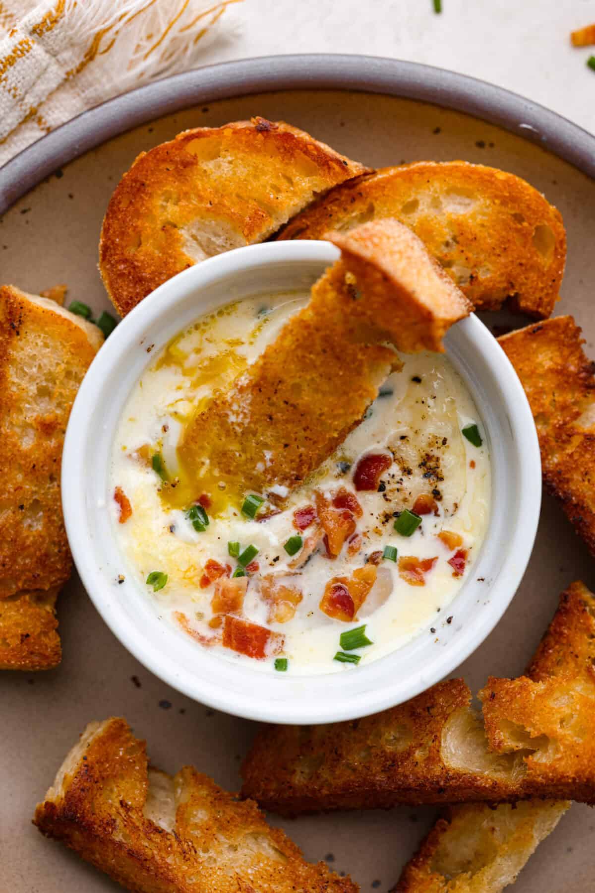 Shirred Eggs are a breakfast worth waking up for in the morning! They're a creamy, custardy combination of eggs, cheese, cream, and bacon that bake in individual ramekins. It's a simple yet super tasty way to start the day! Overhead shot of shirred eggs in a ramekin surrounded by crostini.
