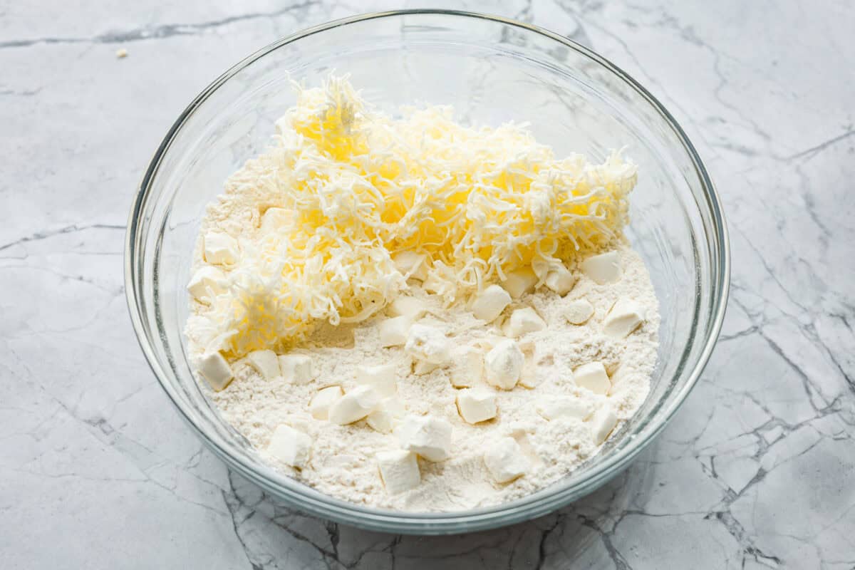 Cream cheese biscuits have a slight twist to the classic version. With the added cream cheese, they have a softer, more fluffy texture with an added tangy flavor that is so good! Overhead shot of flour mixture with shredded butter and cubes of cream cheese in a bowl.
