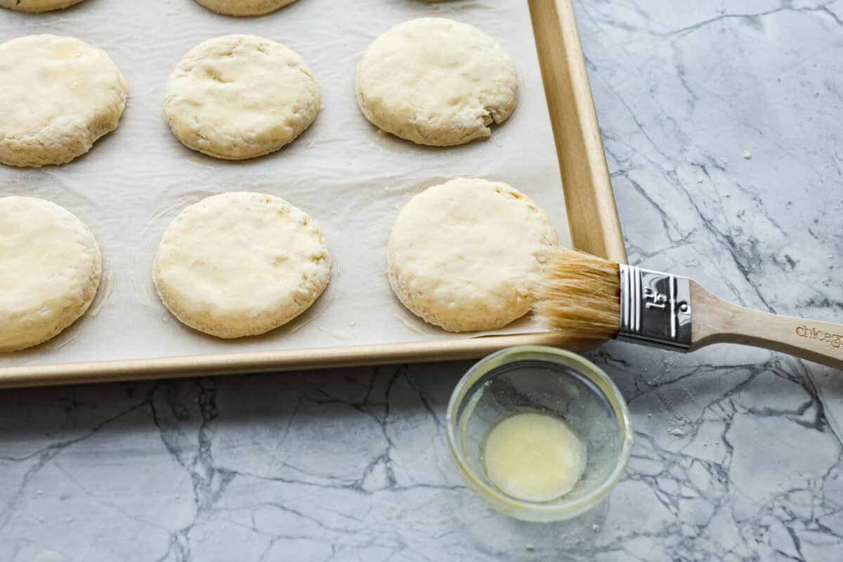 Cream cheese biscuits have a slight twist to the classic version. With the added cream cheese, they have a softer, more fluffy texture with an added tangy flavor that is so good! Overhead shot of biscuit round being brushed with butter before being baked.