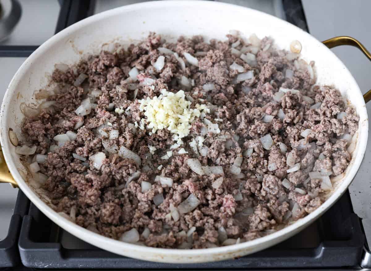 Hamburger Potato Casserole is hearty, filling, and great for family dinners. It's full of tender potatoes and ground beef, smothered in a deliciously creamy sauce, and covered in melty cheese. Angle shot of cooked ground beef and onions in a skillet with garlic and seasonings over the top.