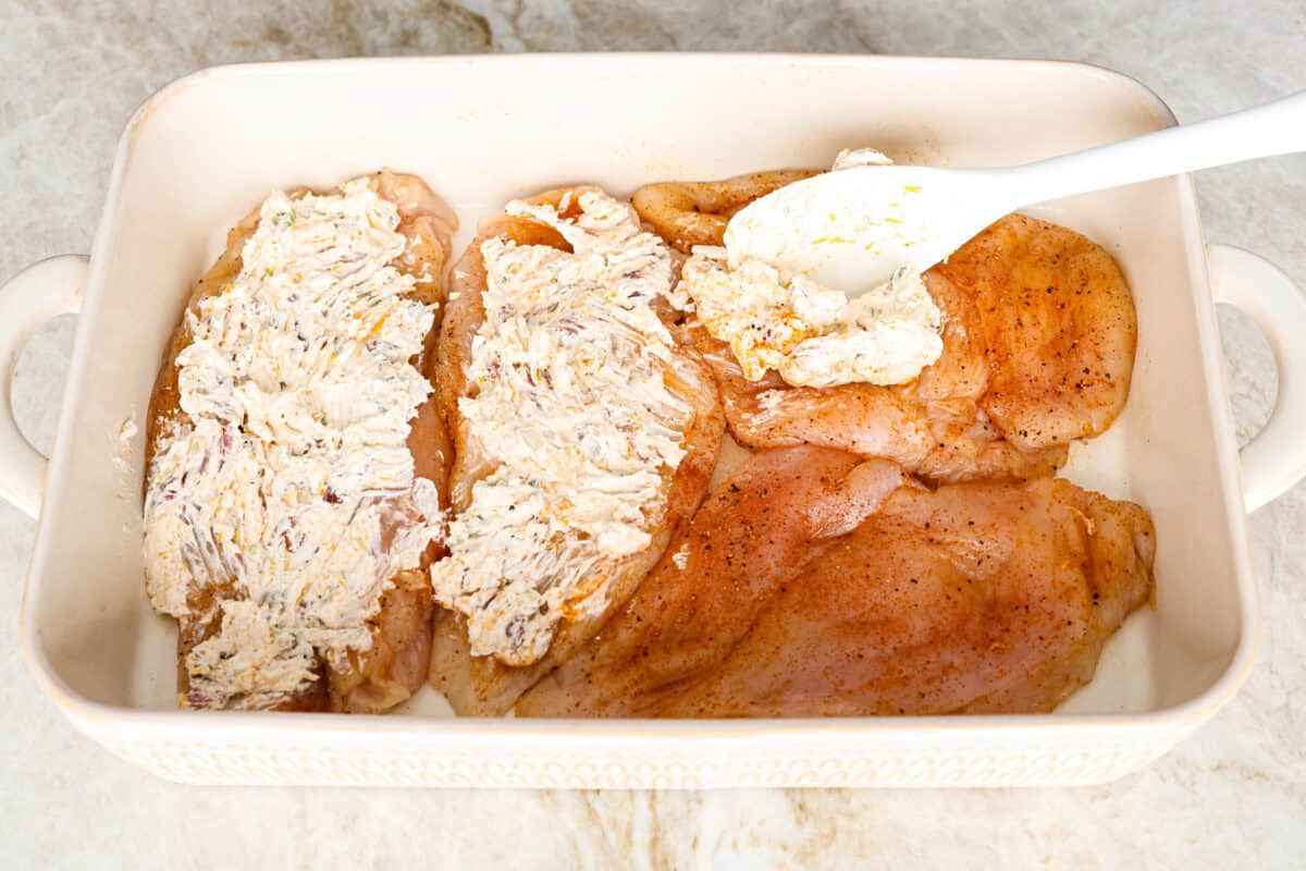 Million Dollar Chicken features juicy baked chicken topped with a creamy, cheesy layer that’s bursting with flavor. The crispy bacon and crunchy almonds really take this to the next level. Enjoy with rice, pasta, vegetables, or any of your favorite sides! Angle shot of someone covering the seasoned chicken breasts with the cream cheese topping mixture in a baking dish.