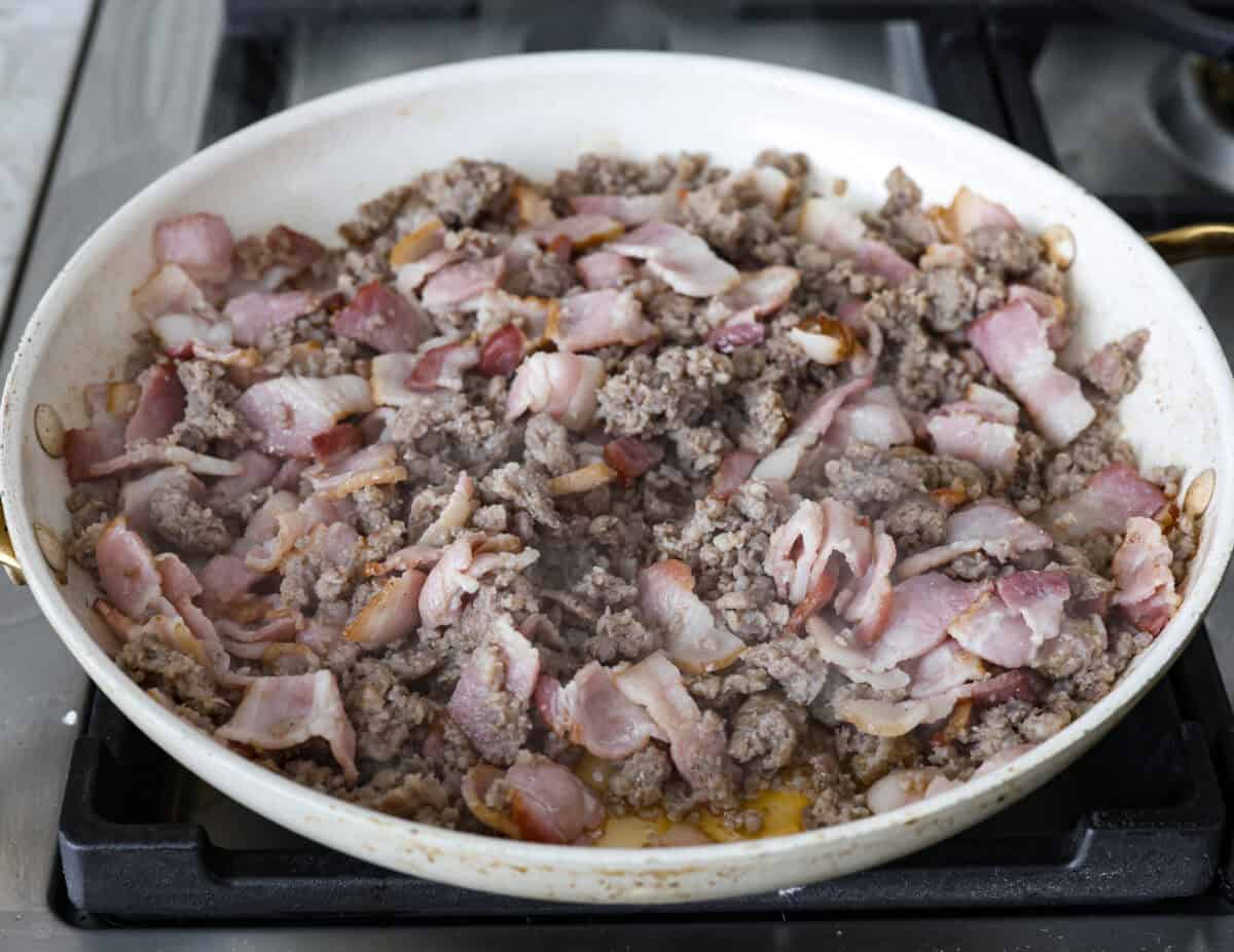 Crack breakfast casserole gets its name for a reason. It is bursting with delicious flavors you won't want to stop eating! It's packed with layers of shredded potatoes, crispy bacon, sausage, and smooth cream cheese, topped with a layer of crispy cheese. Angle shot of bacon and sausage being cooked in a skillet.