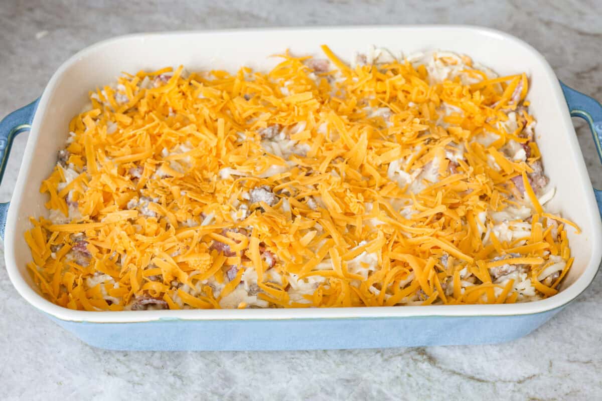 Crack breakfast casserole gets its name for a reason. It is bursting with delicious flavors you won't want to stop eating! It's packed with layers of shredded potatoes, crispy bacon, sausage, and smooth cream cheese, topped with a layer of crispy cheese. Angle shot of casserole ingredients in a baking dish covered with cheese ready to go into the oven.