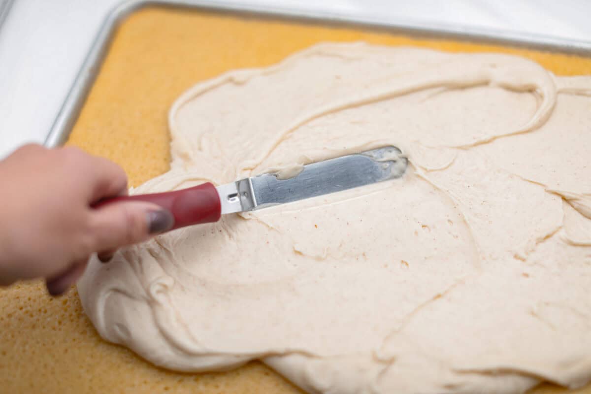 Browned Butter Sheet Cake is the perfect blend of moist, sweet, and irresistibly nutty. With the rich, caramel-like flavor of browned butter baked into every bite, this cake is a treat for anyone who loves a touch of warmth in their desserts. Angle shot of someone spreading the frosting over the cooled sheet cake.