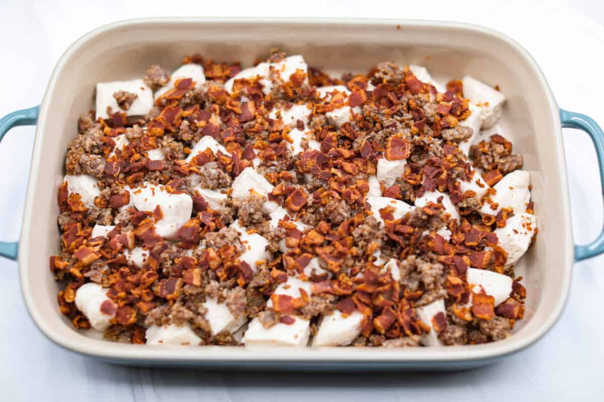 This bubble up breakfast casserole is simple to prepare and super flavorful! It's made with layers of biscuit, crispy bacon, savory sausage, fluffy eggs and melty cheese. Everyone will get out of bed and run to the breakfast table for this yummy dish! Overhead shot of bacon and sausage spread out over the biscuit pieces in the baking dish.