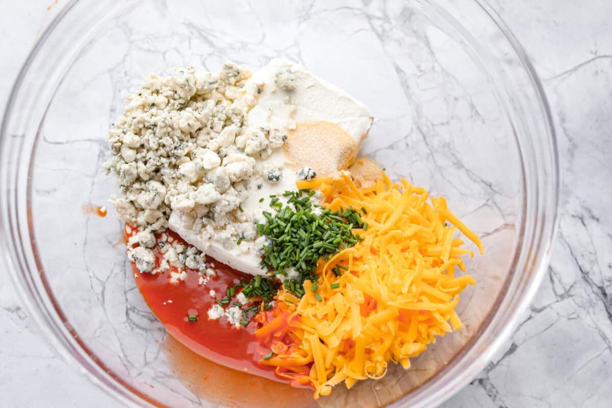 These zesty Buffalo Chicken Pinwheels are an easy snack or appetizer that everyone will devour! A creamy buffalo-flavored spread full of shredded chicken, seasonings, and cheeses all rolled inside of a soft tortilla. Slice into cute pinwheels and enjoy! First photo of the buffalo cream cheese ingredients in a glass bowl.