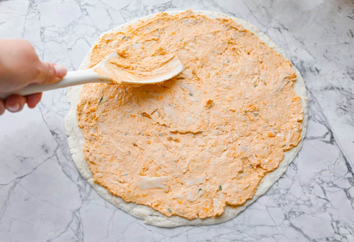 These zesty Buffalo Chicken Pinwheels are an easy snack or appetizer that everyone will devour! A creamy buffalo-flavored spread full of shredded chicken, seasonings, and cheeses all rolled inside of a soft tortilla. Slice into cute pinwheels and enjoy! Fourth photo of the buffalo chicken mixture spreading onto a tortilla