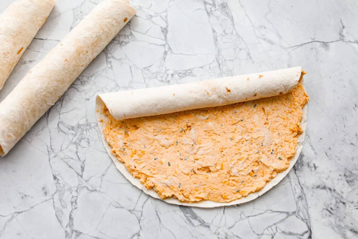 These zesty Buffalo Chicken Pinwheels are an easy snack or appetizer that everyone will devour! A creamy buffalo-flavored spread full of shredded chicken, seasonings, and cheeses all rolled inside of a soft tortilla. Slice into cute pinwheels and enjoy! Fifth photo of the tortilla rolling.