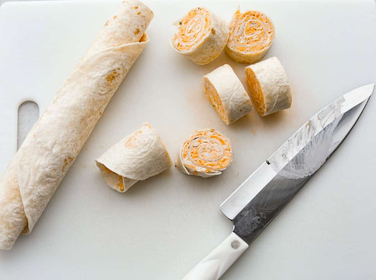 These zesty Buffalo Chicken Pinwheels are an easy snack or appetizer that everyone will devour! A creamy buffalo-flavored spread full of shredded chicken, seasonings, and cheeses all rolled inside of a soft tortilla. Slice into cute pinwheels and enjoy! Sixth photo of the pinwheels being sliced.
