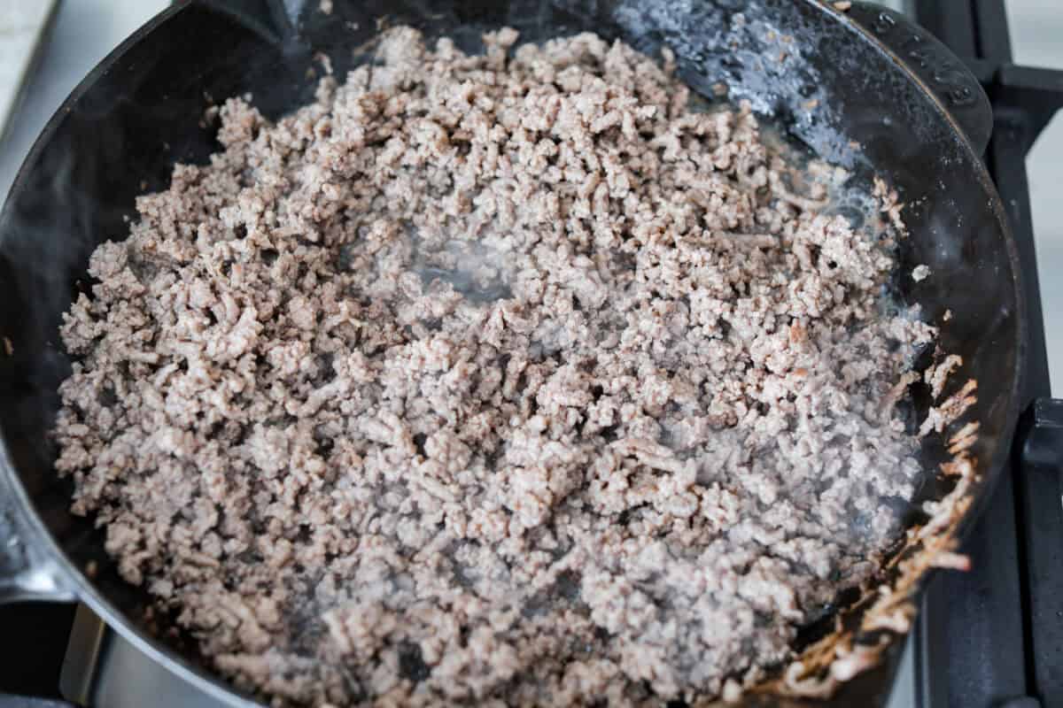 Cheeseburger dip is a crowd-pleasing appetizer for potlucks, parties, and get-togethers! This recipe takes delicious cheeseburger ingredients like beef, onion, tomato, relish, and cheese and turns them into a deliciously addicting snack! Overhead shot of beef being browned in a skillet.