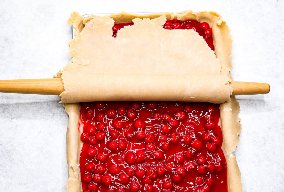 Cherry Slab Pie is an American classic made to serve a crowd! This recipe uses a tender, flaky crust that surrounds sweet, juicy cherry pie filling. It tastes incredibly delicious and is the perfect dessert for any time of year. Overhead shot of the sheet pan with pie crust, pie filling and another layer of pie crust being rolled over the top.