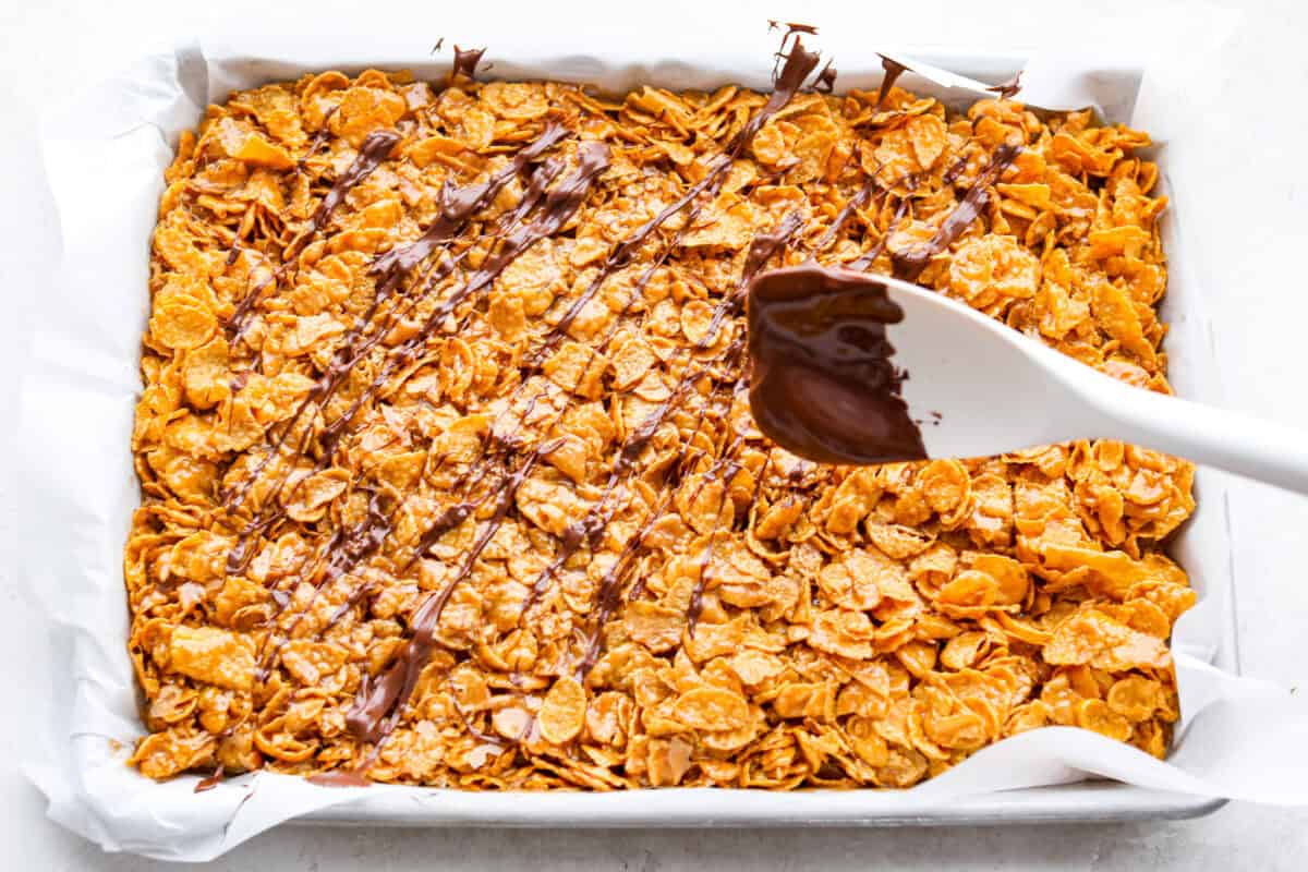 This cornflake brittle crack is crispy, chewy and to die for! With only six ingredients and 8 minutes of prep, you can have this yummy treat ready in no time. This brittle is crunchy cornflakes mixed with a buttery caramel sauce and drizzled in rich dark chocolate. Overhead shot of someone drizzling chocolate over the cornflake and caramel mixture.