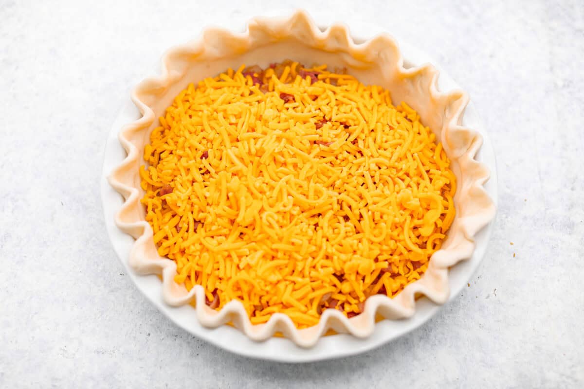 Cowboy Quiche is the ultimate hearty, satisfying breakfast that brings classic comfort to the table. It is filled with smoky bacon, caramelized onions, and cheddar cheese, all baked together in a buttery pie crust. It’s rich, savory, and packed with bold flavor. Overhead shot of bacon onions and cheese in the bottom of the pie crust.