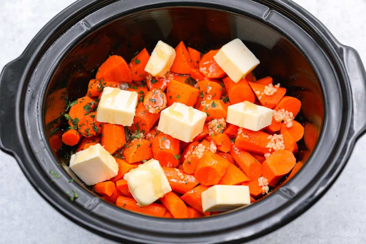 These crockpot carrots will become fork-tender from slowly cooking in your crockpot all day. The carrots have a delicious sweet and savory flavor from the sticky honey and bold garlic, all slathered in butter. They are the perfect side for your next Thanksgiving or Easter feast. Overhead shot of carrots with honey, butter, garlic, parsley, salt and pepper in a crockpot.
