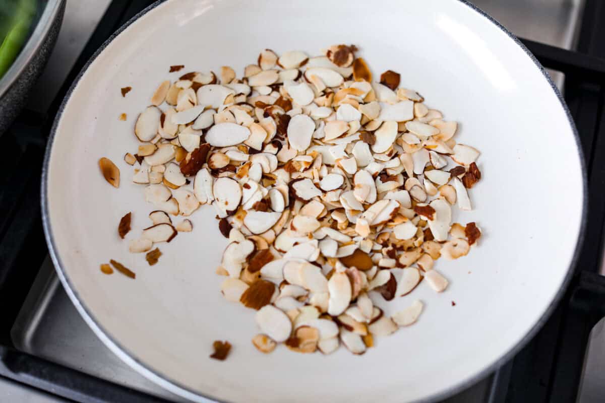 Green Beans Almondine is made with fresh green beans that are sautรฉed until crisp-tender and then tossed with buttery, toasted almonds. Every bite is filled with nutty richness and a hint of lemon to brighten up the flavors. Overhead shot of almonds toasting in a skillet.