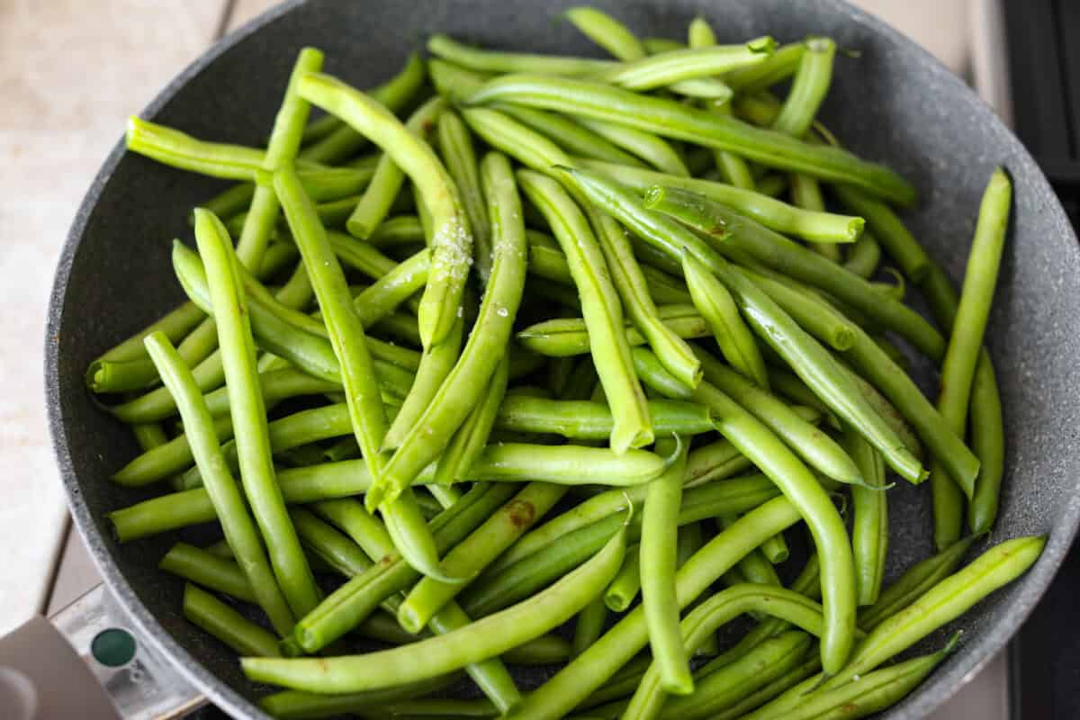 Green Beans Almondine is made with fresh green beans that are sautรฉed until crisp-tender and then tossed with buttery, toasted almonds. Every bite is filled with nutty richness and a hint of lemon to brighten up the flavors. Overhead shot of green beans, salt and water in a skillet.