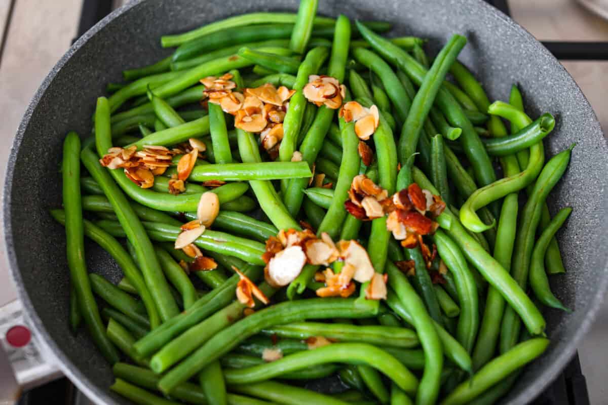 Green Beans Almondine is made with fresh green beans that are sautรฉed until crisp-tender and then tossed with buttery, toasted almonds. Every bite is filled with nutty richness and a hint of lemon to brighten up the flavors. Overhead shot of steamed green beans with the almond, butter and lemon juice poured over the top.
