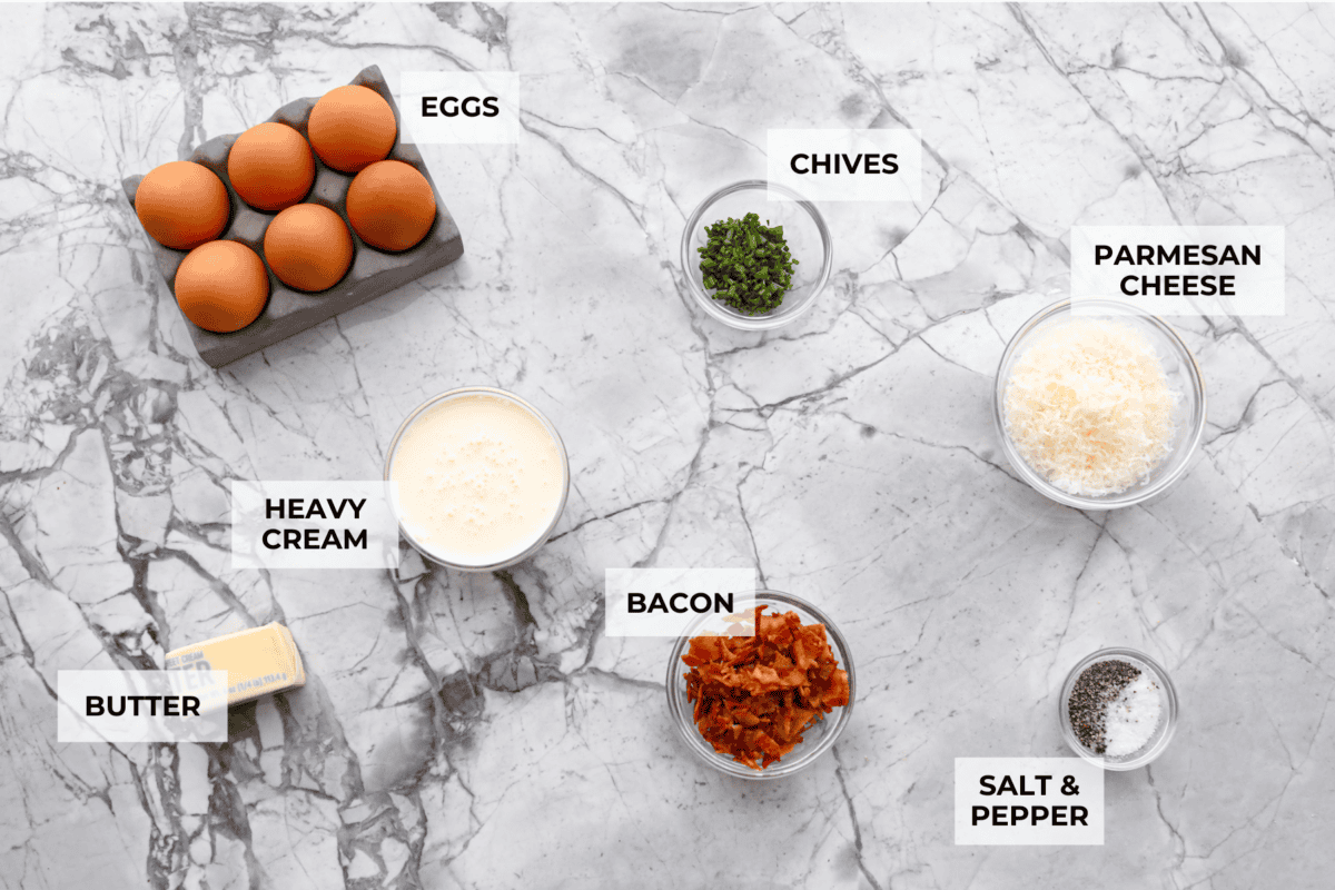 Shirred Eggs are a breakfast worth waking up for in the morning! They're a creamy, custardy combination of eggs, cheese, cream, and bacon that bake in individual ramekins. It's a simple yet super tasty way to start the day! Overhead shot of labeled ingredients.