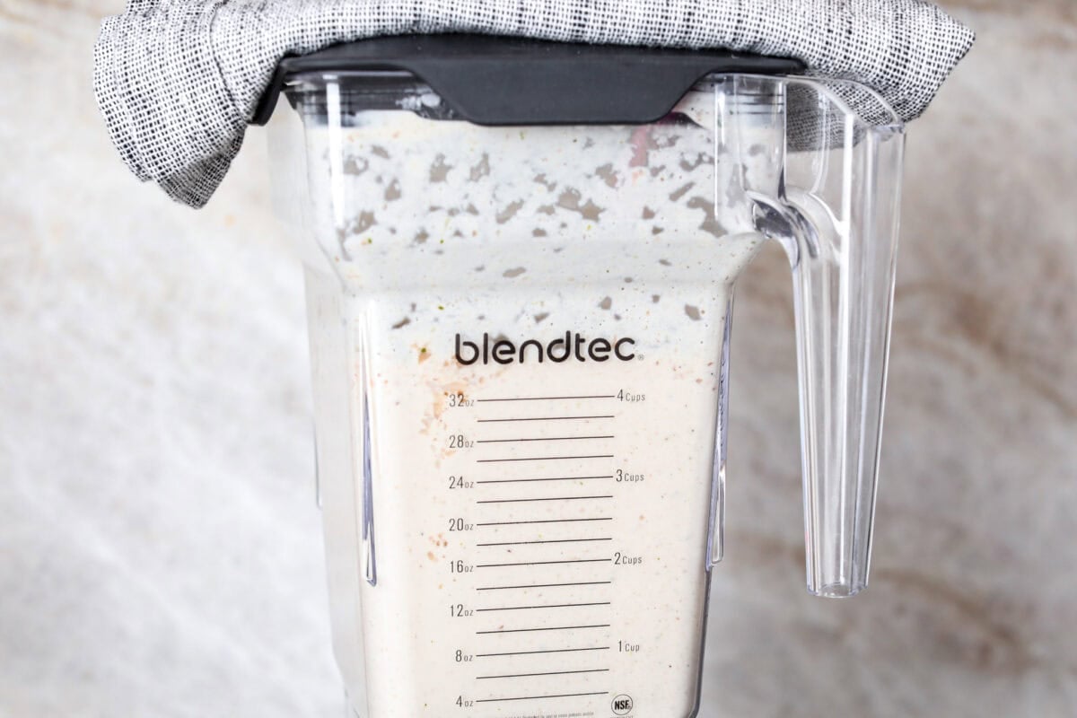 Mushroom bisque is full of cream and garlic-infused mushrooms, all blended to make a thick, rich, and beyond flavorful soup! You will want to drink this straight from your bowl! Side shot of blender with soup ingredients inside with towel on top.