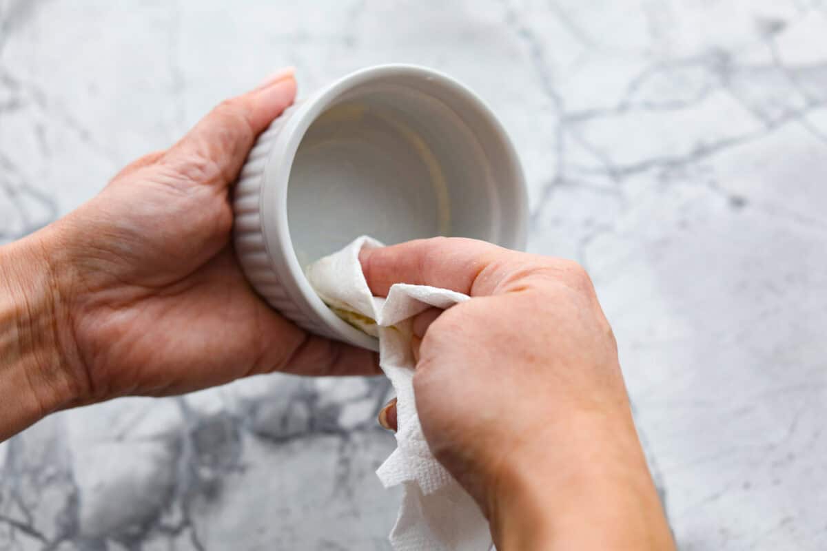 Shirred Eggs are a breakfast worth waking up for in the morning! They're a creamy, custardy combination of eggs, cheese, cream, and bacon that bake in individual ramekins. It's a simple yet super tasty way to start the day! Overhead shot of someone prepping a ramekin with butter.