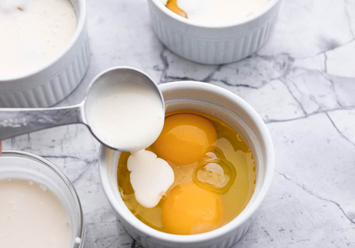Shirred Eggs are a breakfast worth waking up for in the morning! They're a creamy, custardy combination of eggs, cheese, cream, and bacon that bake in individual ramekins. It's a simple yet super tasty way to start the day! Overhead shot of ramekin with two eggs and someone pouring cream in it.
