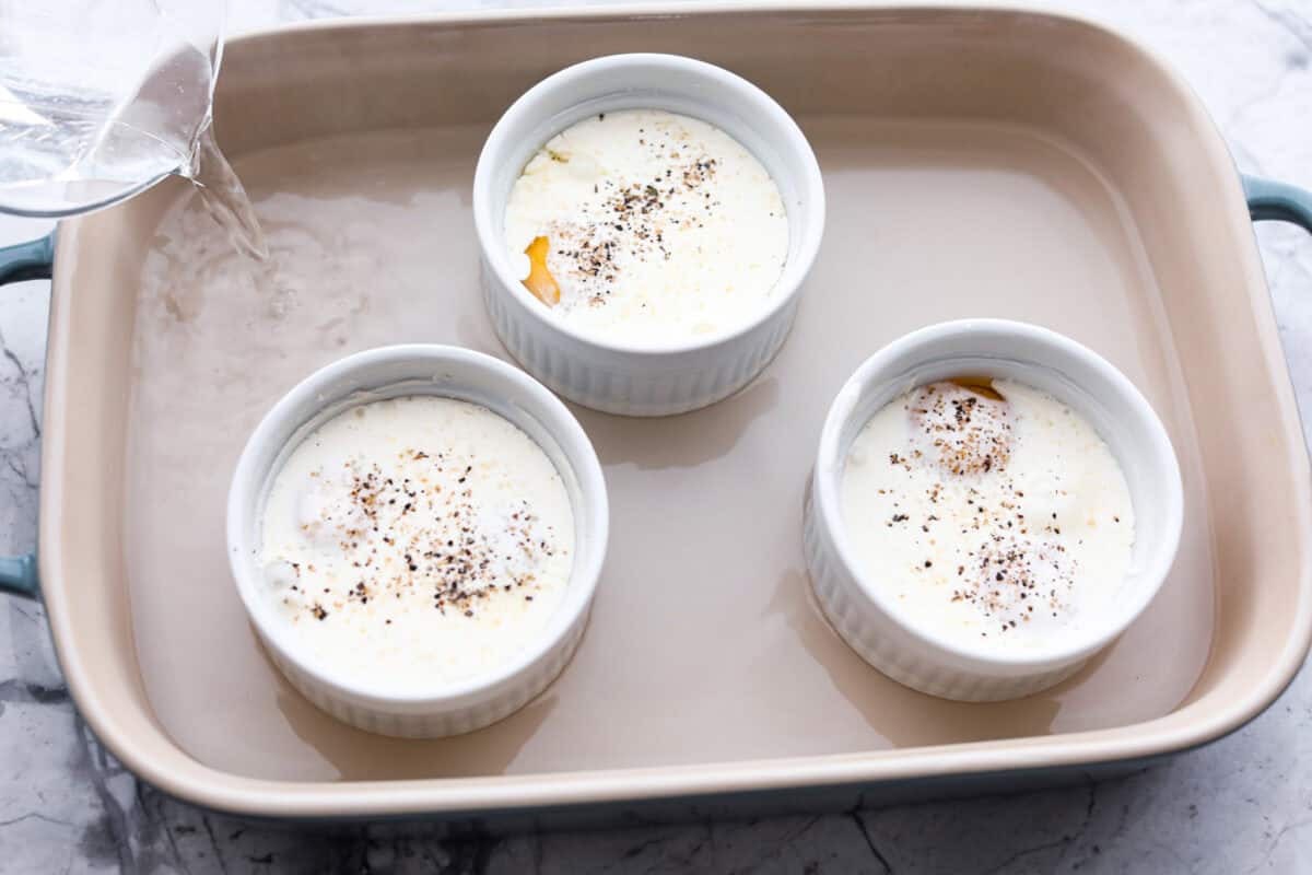Shirred Eggs are a breakfast worth waking up for in the morning! They're a creamy, custardy combination of eggs, cheese, cream, and bacon that bake in individual ramekins. It's a simple yet super tasty way to start the day! Overhead shot of three ramekins in baking dish with water being poured into the baking dish surrounding the ramekins.