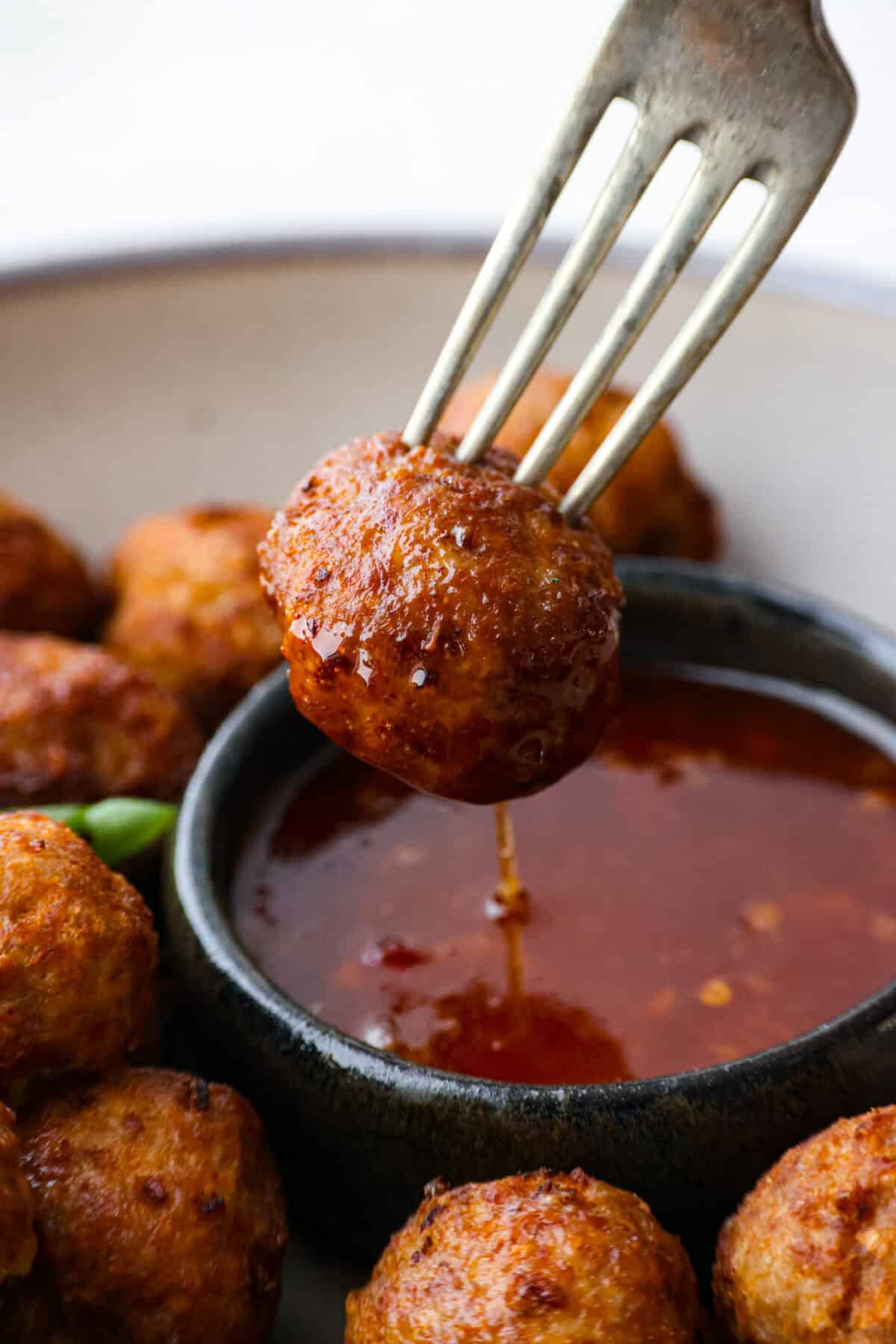 Easy Bola Bola (Filipino meatballs) combine pork, panko breadcrumbs, carrots, and onions to create a versatile meatball recipe. Tender and tasty on the inside then deep-fried and crispy on the outside! The perfect appetizer or dinner idea! Close view of a fork dipping a meatball into a bowl of sweet chili sauce.