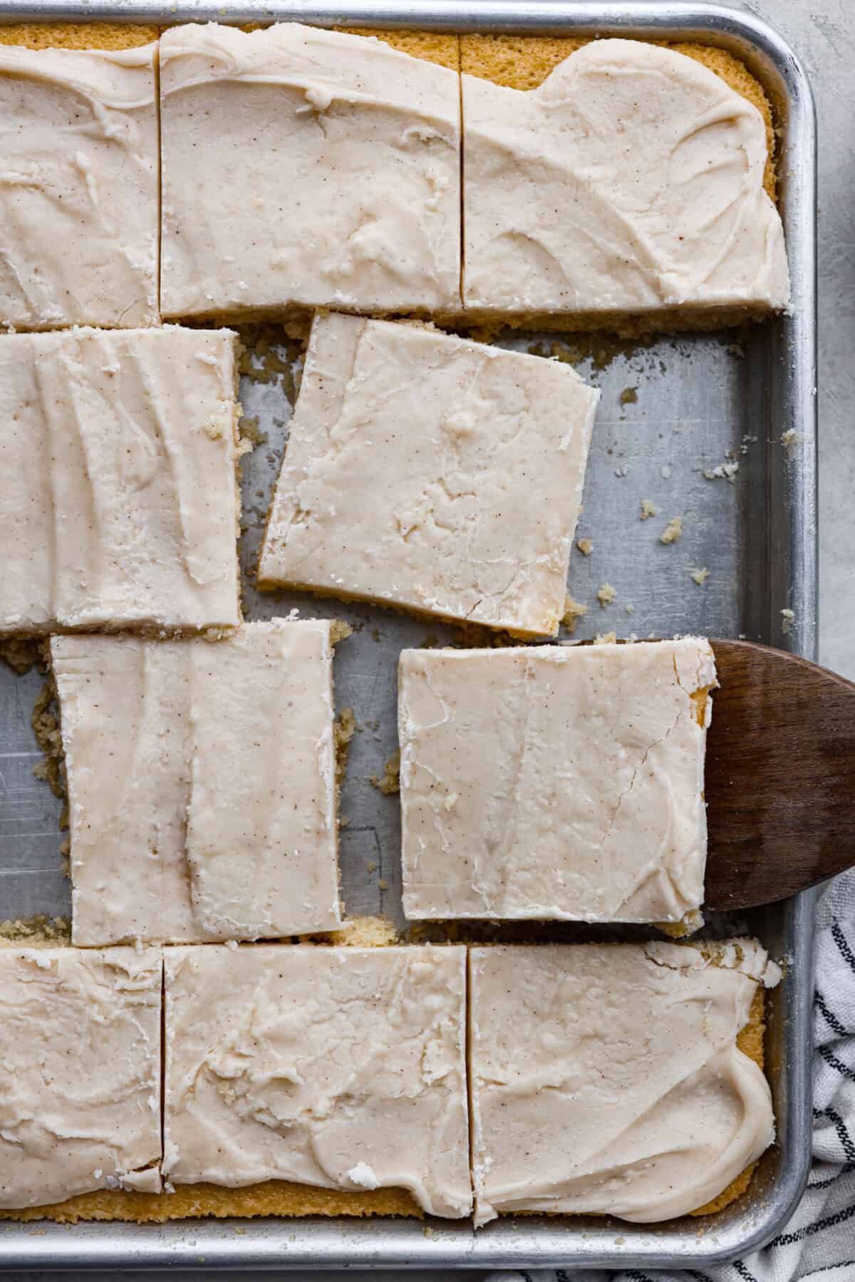 Browned Butter Sheet Cake is the perfect blend of moist, sweet, and irresistibly nutty. With the rich, caramel-like flavor of browned butter baked into every bite, this cake is a treat for anyone who loves a touch of warmth in their desserts. Overhead shot of browned butter sheet cake cut into squares in the baking sheet with someone taking one of the squares out with a wooden spatula.