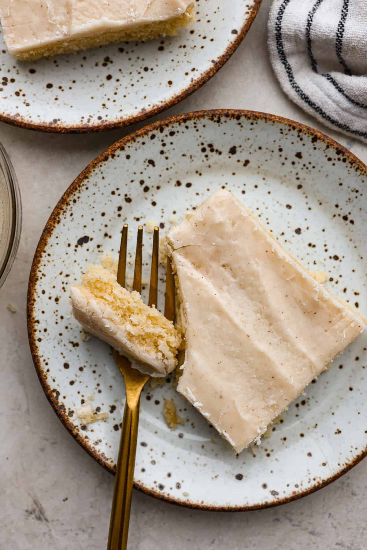Browned Butter Sheet Cake is the perfect blend of moist, sweet, and irresistibly nutty. With the rich, caramel-like flavor of browned butter baked into every bite, this cake is a treat for anyone who loves a touch of warmth in their desserts. Close up shot of a plated peice of browned butter sheet cake with a bite taken out with a gold fork.