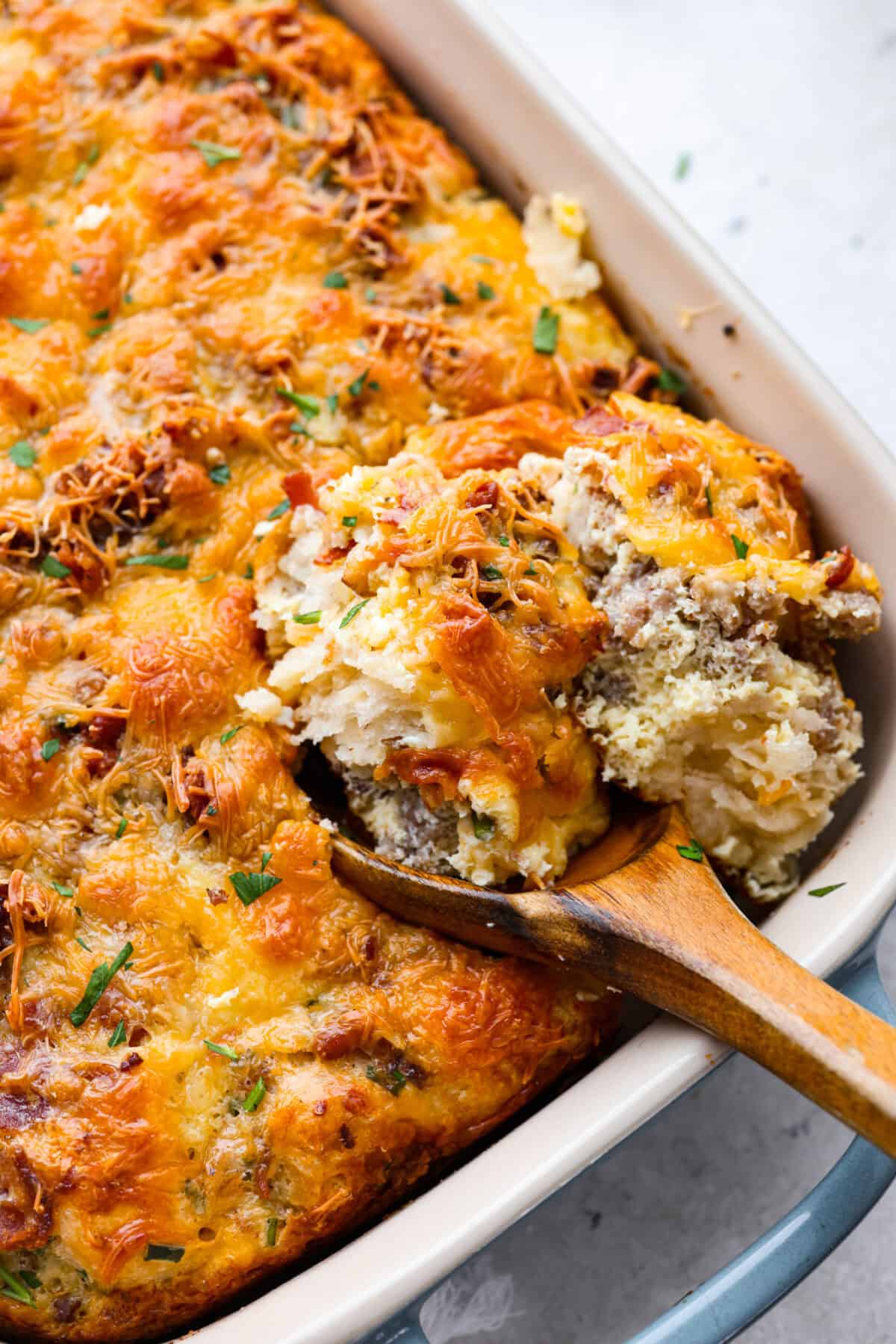 This bubble up breakfast casserole is simple to prepare and super flavorful! It's made with layers of biscuit, crispy bacon, savory sausage, fluffy eggs and melty cheese. Everyone will get out of bed and run to the breakfast table for this yummy dish! Close up shot of bubble up breakfast casserole in a baking dish with a wooden serving spoon.