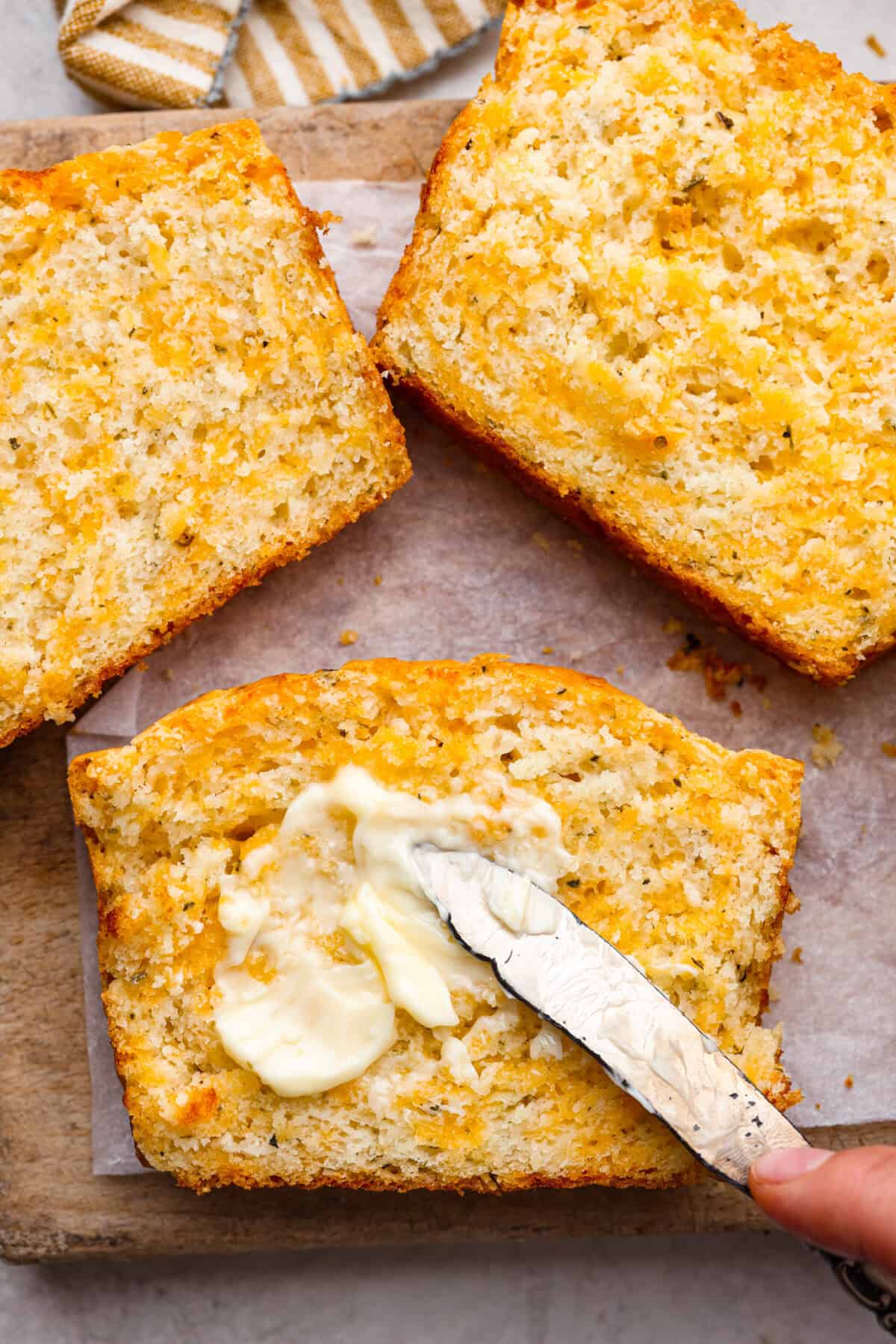Easy Cheesy Quick Bread is packed with flavor and has a tender, golden crust and soft, moist interior. It's perfect with soup, salad, chili, or on its ownย with a pat of butter. It's simple to make and irresistibly delicious! Close up shot of someone spreading butter on a slice of cheesy quick bread.