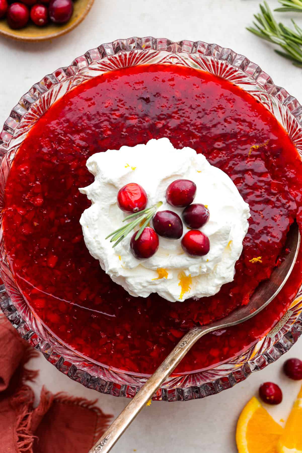 Cranberry Jello Salad is the best combination of tart, tangy, and sweet! It's similar to cranberry sauce but a little sweeter from the jello with undertones of orange and pineapple. Add it to your holiday spread and watch it disappear! Overhead shot of cranberry jello salad with whipped cream on top and whole cranberry garnish.