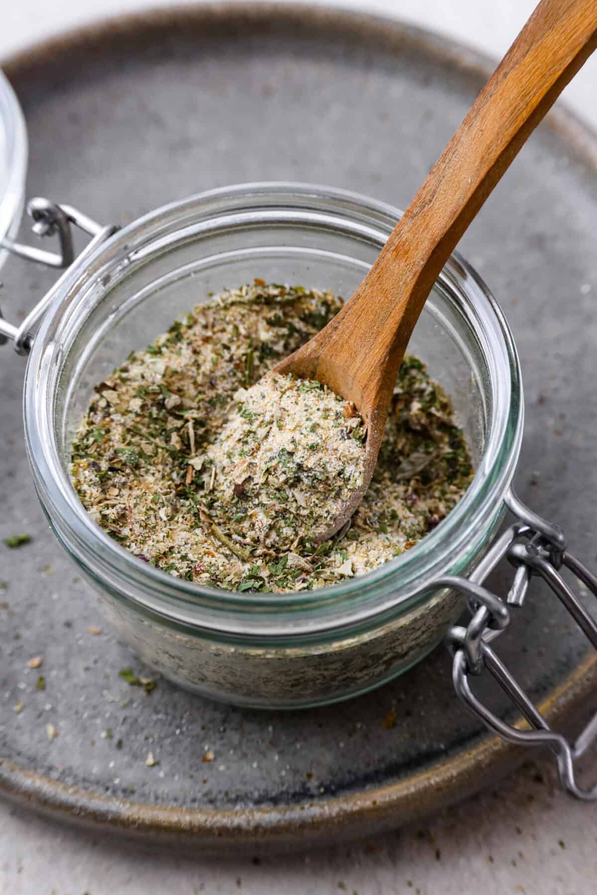 Homemade garlic herb seasoning is a great mix to have on hand. It has multiple uses, is full of flavor, and is adjustable to suit your taste preferences. It has the perfect mix of herbaceous dried herbs, garlic, onion powder, salt, and fresh cracked pepper. Angle shot of homemade garlic herb seasoning in a glass jar with a small wooden spoon sticking out.