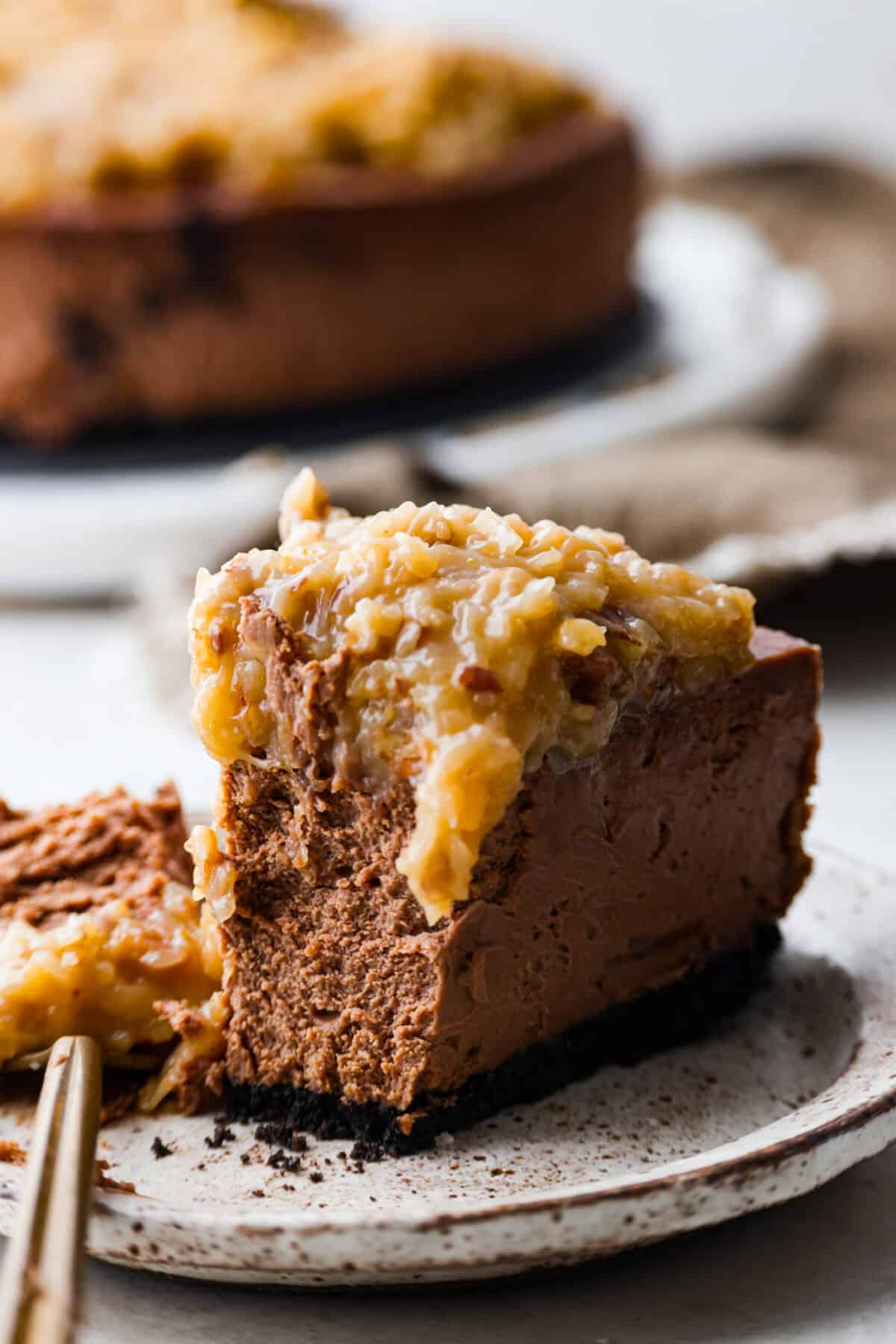 German Chocolate Cheesecake is a rich and impressive dessert! It has an Oreo cookie crust, a chocolate cheesecake filling, and a classic German chocolate topping to finish it off! The gooey coconut pecan topping takes this cheesecake to the next level! Close view of a slice of German chocolate cheesecake on a plate.