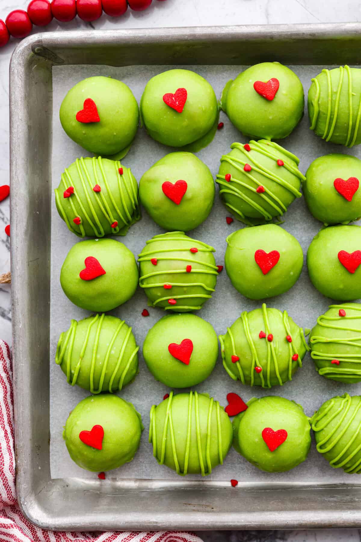 These are the cutest Grinch Oreo balls you have ever seen! Fudgy Oreo filling is coated in green candy melts and garnished with the cutest red heart sprinkle. They taste amazing and will steal the show this holiday season at holiday parties, family get-togethers, or Christmas events. Overhead shot of a cookie sheet full of completed Grinch Oreo balls with red heart sprinkles.