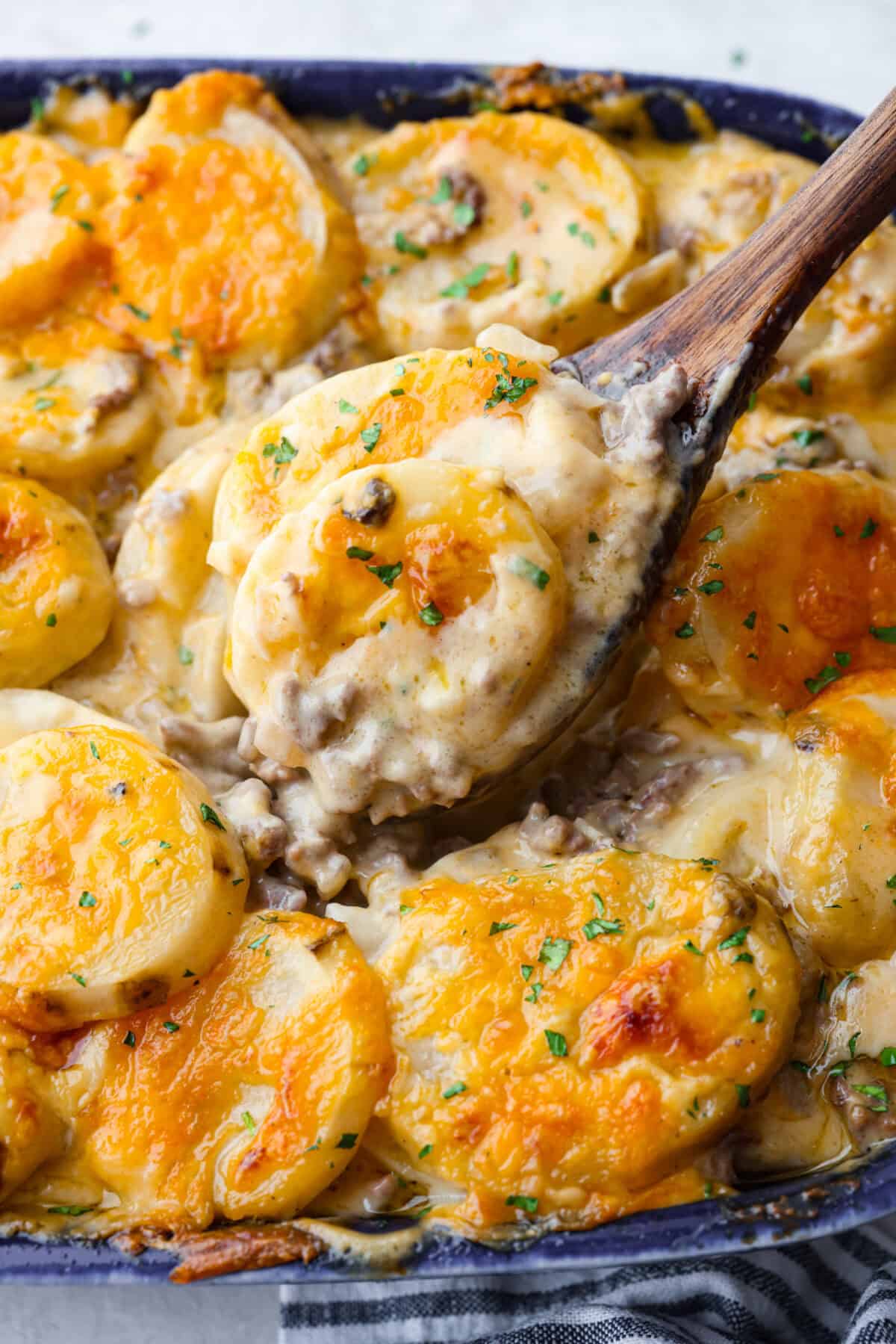 Hamburger Potato Casserole is hearty, filling, and great for family dinners. It's full of tender potatoes and ground beef, smothered in a deliciously creamy sauce, and covered in melty cheese. Close up shot of a scoop of hamburger potato casserole on a wooden spoon.
