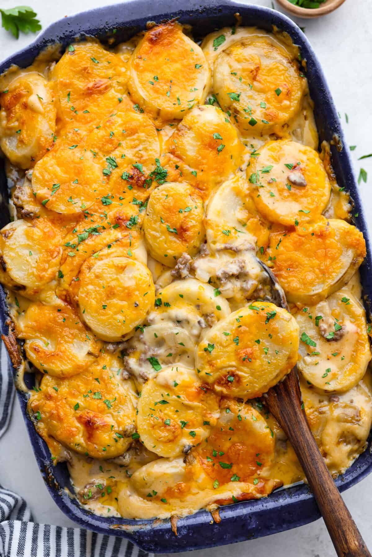 Hamburger Potato Casserole is hearty, filling, and great for family dinners. It's full of tender potatoes and ground beef, smothered in a deliciously creamy sauce, and covered in melty cheese. Overhead shot of hamburger potato casserole with wooden serving spoon.